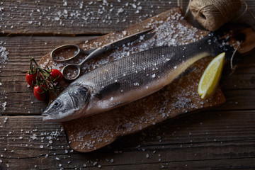 Sea bass fish on the dark wooden background with salt, tomatoes cherry and lemon