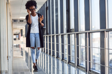 Young african american female passanger in casual clothes is in airport listening music in headphones