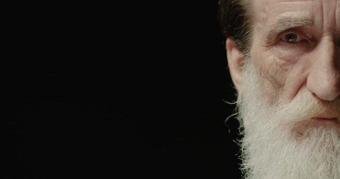 Close up portrait of good-looking Caucasian senior man half face on black background looking with smile on face indoors. Handsome bearded old male model posing to camera and smiling. Emotions concept