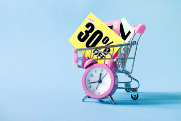 Mini shopping cart with a thirty percent discount sign and pink alarm clock on blue background....