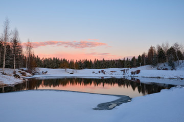 Winter landscape. The forest lake is partially covered with ice, surrounded by rocks, shrubs and spruces against the evening sky painted by the rays of the setting sun.