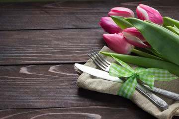  Table setting with pink tulips  and cutlery on dark wooden board.