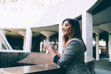 Trendy young female blogger using mobile phone for share multimedia enjoying free time in modern building, happy millennial student woman chatting and sending messages on smartphone on break