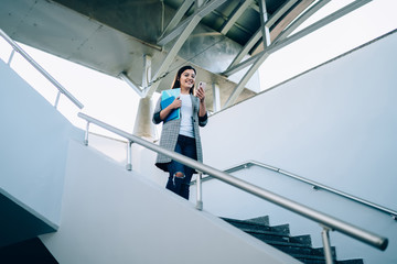 Smiling  young woman reading notification on mobile phone walking down on urban staircase, cheerful trendy dressed millennial hipster girl happy about getting good 4G internet send text message