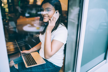 Cheerful caucasian woman in spectacles making cellular call while working remotely on freelance, smiling female student talking on smartphone doing homework on laptop computer in coworking space