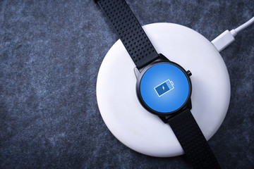 Smart watch on wireless charging with on-screen charging indicator. At the desktop, near the laptop. Top view. Place for text