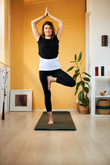 Middle aged brunette standing in Tree yoga pose at home in the morning.