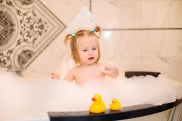 Toddler girl bathes in bath with foam at home, plays with rubber duck