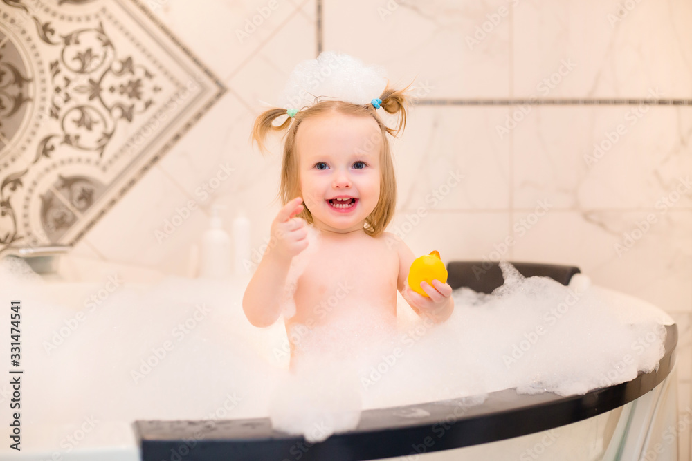 Wall mural Toddler girl bathes in bath with foam at home, plays with rubber duck