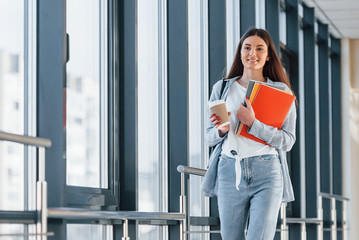 Female young student is in corridor of a college holding notepads and cup of drink