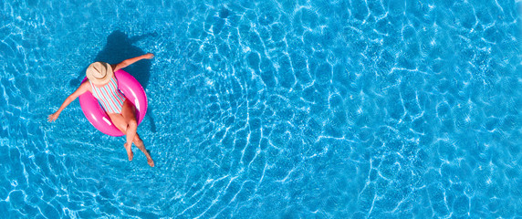 Zenith aerial view of a swimming pool in summer. Young girl in a swimsuit and hat floating with pink donut. - obrazy, fototapety, plakaty