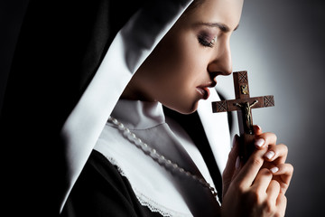 sad young nun praying with cross on grey