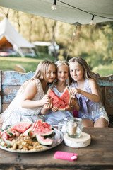 Three cute happy smiling girls, sisters, fiends, sitting at the table on vintage wooden bench and eating watermelon outdoors, in tent, stylish boho wigwam on background. Countryside summer holidays