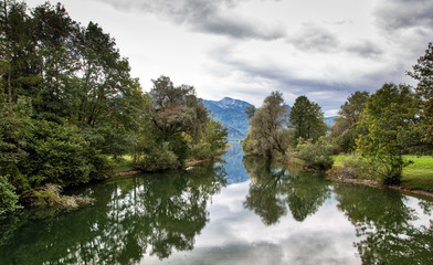 Fototapeta na wymiar landscape panorama of river loisach on the way in the lake Kochel with trees and reflections of the trees in the water. In the background the lake kochel and the mountains of the alps