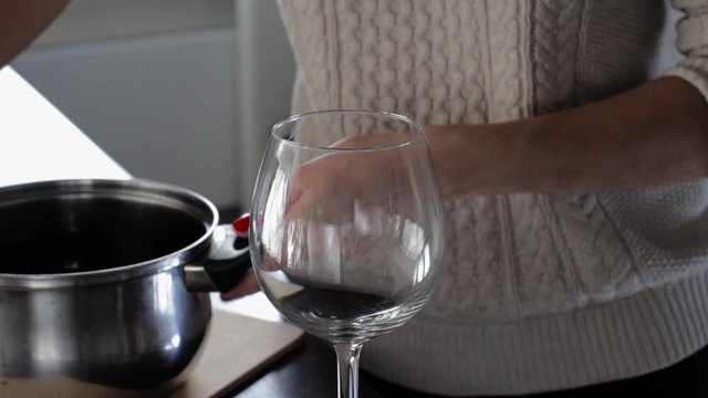 young woman wearing a beige sweater and making mulled wine with spices in a kitchen. woman pouring mulled wine from a pan in a glass