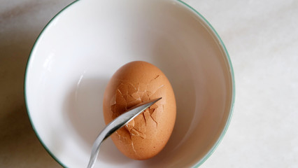 A brown egg inside a bowl, with a silver spoon hitting it and causing cracks on the egg shell. 