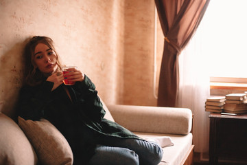 Young bored woman having tea while sitting on sofa in living room at home