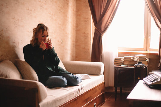Young thoughtful woman drinking tea while sitting on sofa in living room at home