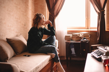 Thoughtful young woman looking through window while sitting on bed at home