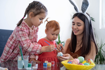 Obraz na płótnie Canvas Happy young beautiful mother spending time with her joyful daughters. Cheerful family mom and children painting easter eggs with colors. Little pretty girl putting bunny ears. Preparation for Easter.