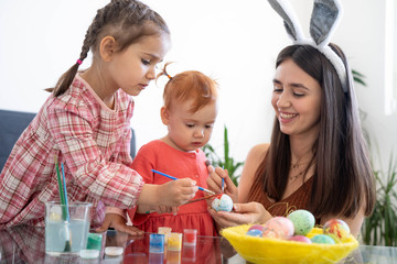 Happy young beautiful mother spending time with her joyful daughters. Cheerful family mom and children painting easter eggs with colors. Little pretty girl putting bunny ears. Preparation for Easter.