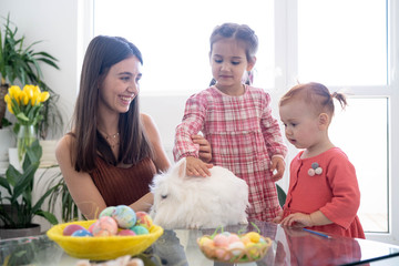 Cheerful smiley mother and her daughters playing with Easter white rabbit and laughing. Happy family preparing for Easter. Easter family concept. White window background. Yellow flowers.