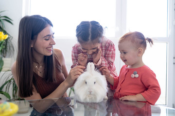 Cheerful smiley mother and her daughters playing with Easter white rabbit and laughing. Happy family preparing for Easter. Easter family concept. White window background. Yellow flowers.