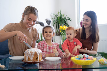 Two beautiful mothers with their daughters celebrating Easter eating cake and having fun together on a sunny morning. Girls having breakfast in a white dining room with a big window. Easter concept.