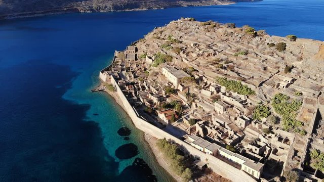 Scenic aerial view of Spinalonga fortress isle, old buildings and structures ruins on rocky slope, shallow sea water at North shore. Popular sight of Crete Island
