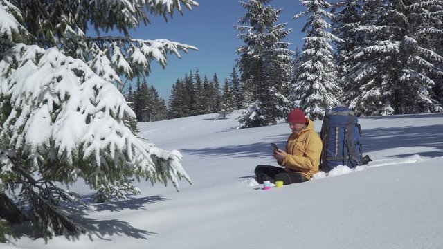 Travaler sits in the snow and talks on the phone in the winter forest. Carpathian mountain range. 4K