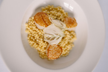 a plate with ptitim pasta, grilled scallops, cream sauce and dried onion slices. Smooth image with shallow depth of field.