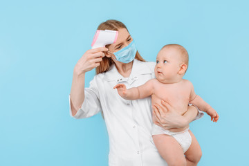 a female pediatrician in a medical mask with a digital infrared thermometer, measures the body...