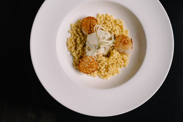 a plate with ptitim pasta, grilled scallops, cream sauce and dried onion slices. Dark background. Smooth image with shallow depth of field.