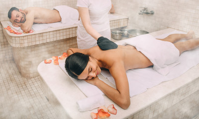 Mixed race woman having skin peeling in turkish hammam while her boyfriend enjoys the warmth of a Turkish bath marble massage table - obrazy, fototapety, plakaty