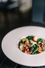 Octopus salad with stracciatella, tomatoes, olives and green basilisk on a plate. Dark table background. Smooth image with shallow depth of field.