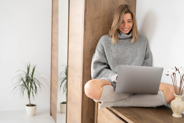 Smiling attractive young woman sitting on wooden furniture using laptop communicating working online at home. Happy cute girl in warm sweater and socks typing on computer. Cozy concept