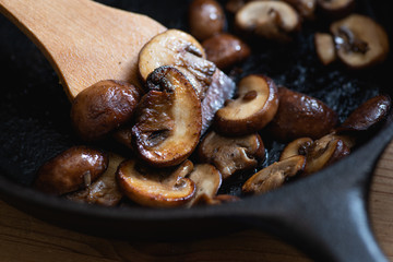 Brown mushrooms cooked in a cast iron skillet, wooden spatula.