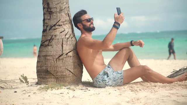 Selfie On Smartphone In Beautiful Place.Man Using Mobile Phone For Selfie Photo.Attractive Tanned Man Sunbathing Using Mobile App For Photo Video.Guy Taking Picture On Vacation Holiday Carribean Beach