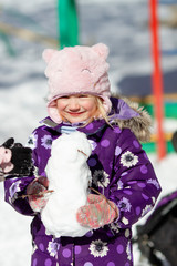 Portrait of happy smiling pretty toddler girl wearing bright colored snowsuit and pink hat holding snowman in a sunny winter day