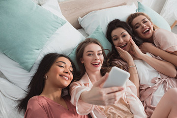 Obraz premium High angle view of excited multicultural women taking selfie while resting on bed at bachelorette party