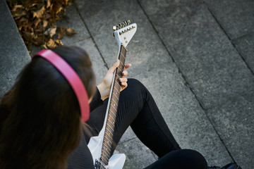 young woman dressed in a sweater and black trousers and playing a white electric guitar sitting on stone stairs and listening with pink headphones