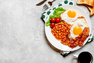 Fried eggs, crispy bacon, beans and tomatoes in a white plate on a gray concrete background, top view, copy space. Breakfast with eggs, bacon and coffee, free space for text.