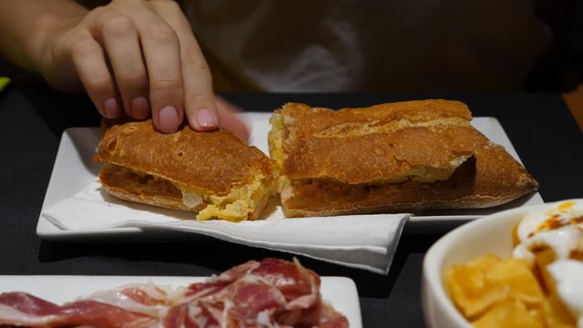 Woman try baguette with potato omelet, meal served at Spanish restaurant. Take half of sandwich and bite it, put back on the plate.