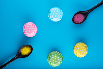 Black spoon. coloured decorative sugar..Muffin case. Pink cupcake liners. Pink Sugar. Cake cases.Top View. Flat laying. Blue background. View from above..Space for text. Blue background. Yellow Sugar