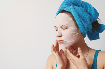 a young beautiful woman uses a moisturizing cosmetic fabric face mask with a towel wrapped around...