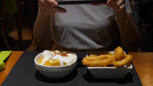Woman using phone to take pictures of tapas, fried squid rings and Patatas bravas with mayo, typical Spanish snacks served in small bowl plates. Tourist girl have brunch or lunch at cafe of Barcelona