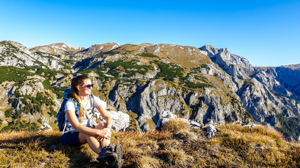 Naklejka na ściany i meble A woman sitting at the top of the Alpine Buchbergkogel, Austria. The woman has a big hiking backpack. She is enjoying the landscape and beautiful day. Exploring new places. Freedom and happiness.