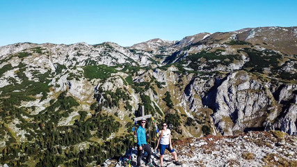 A drone shot of a couple in hiking outfits standing next to a cross on top of Buchbergkogel, Hochschwab region, Austria. They are surrounded by mountains. Achieving a goal. Alpine landscape.