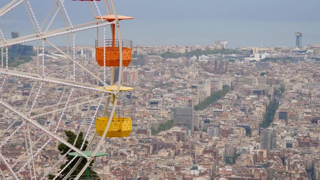 Empty multi-coloured pods of vintage Ferris Wheel carousel move against blurred Barcelona city. Telephoto perspective from height of mountain. L'Eixample district seen on back, urban city sea shore
