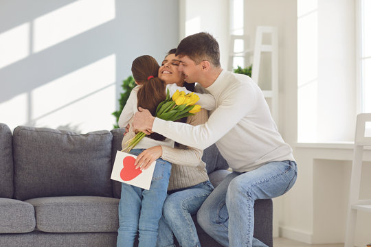 Happy mother's day. Father and baby daughter congratulates mom with flowers and a postcard in a room